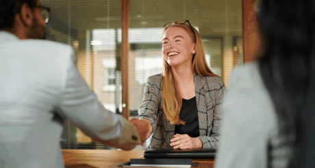 A woman with red hair smiles and shakes the hand of someone out of picture, who is sitting across the table.